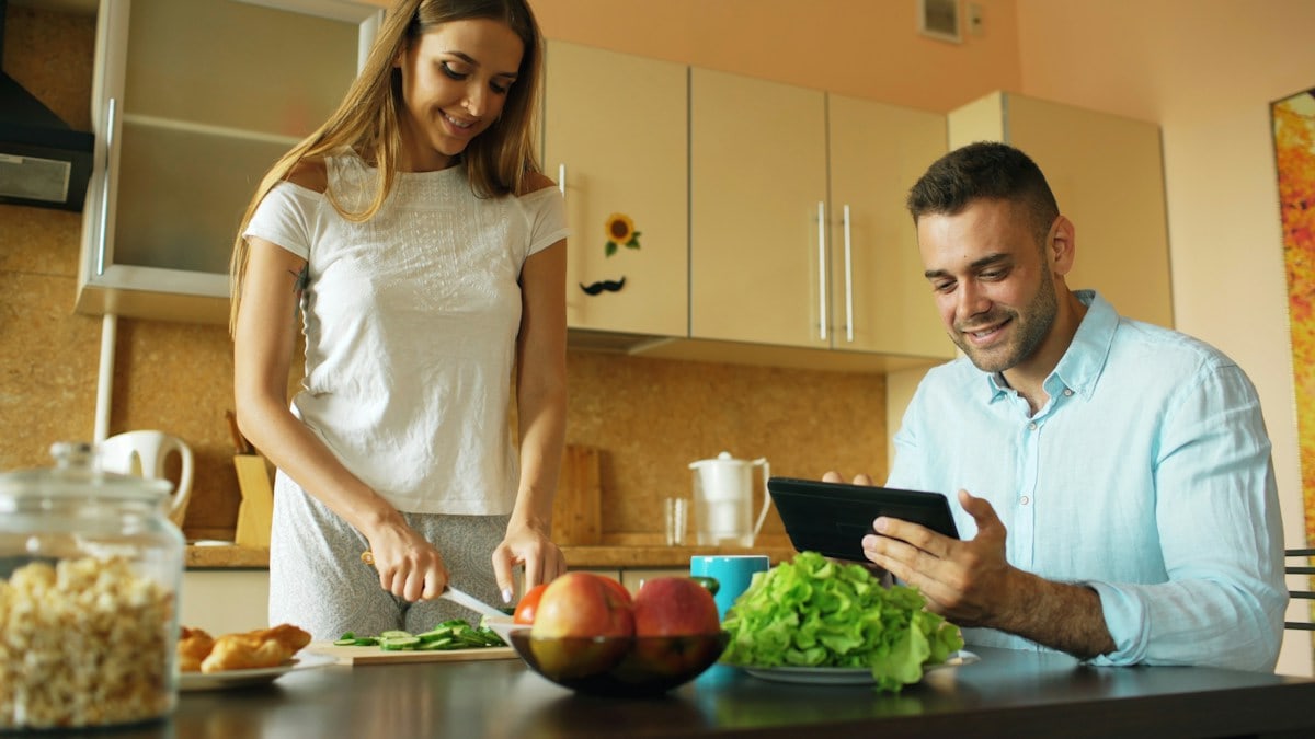 🎬 FULL 4K VIDEO for this still is in my bio link! ⬇️ Attractive couple chatting in the kitchen early morning. Handsome man using tablet while his girlfriend cooking breakfast at home